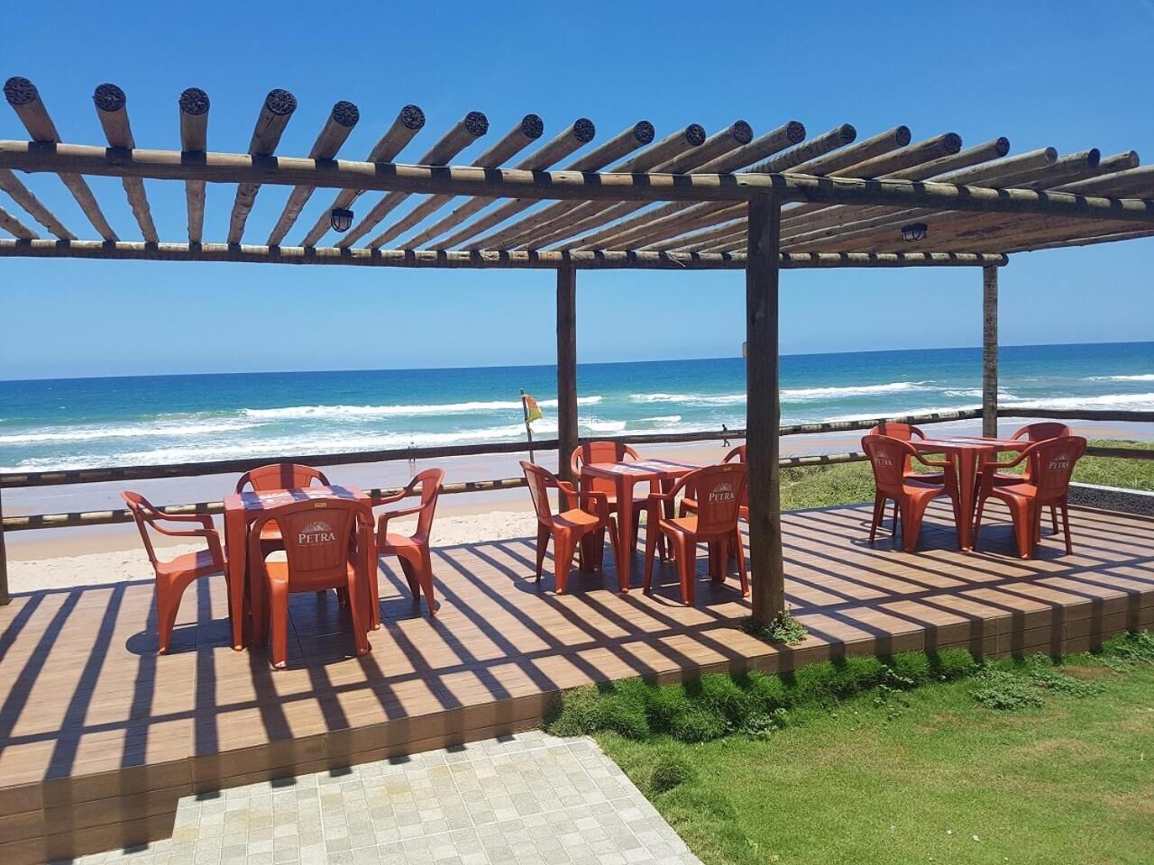 A beachside patio with wooden pergola, red plastic chairs, and tables.