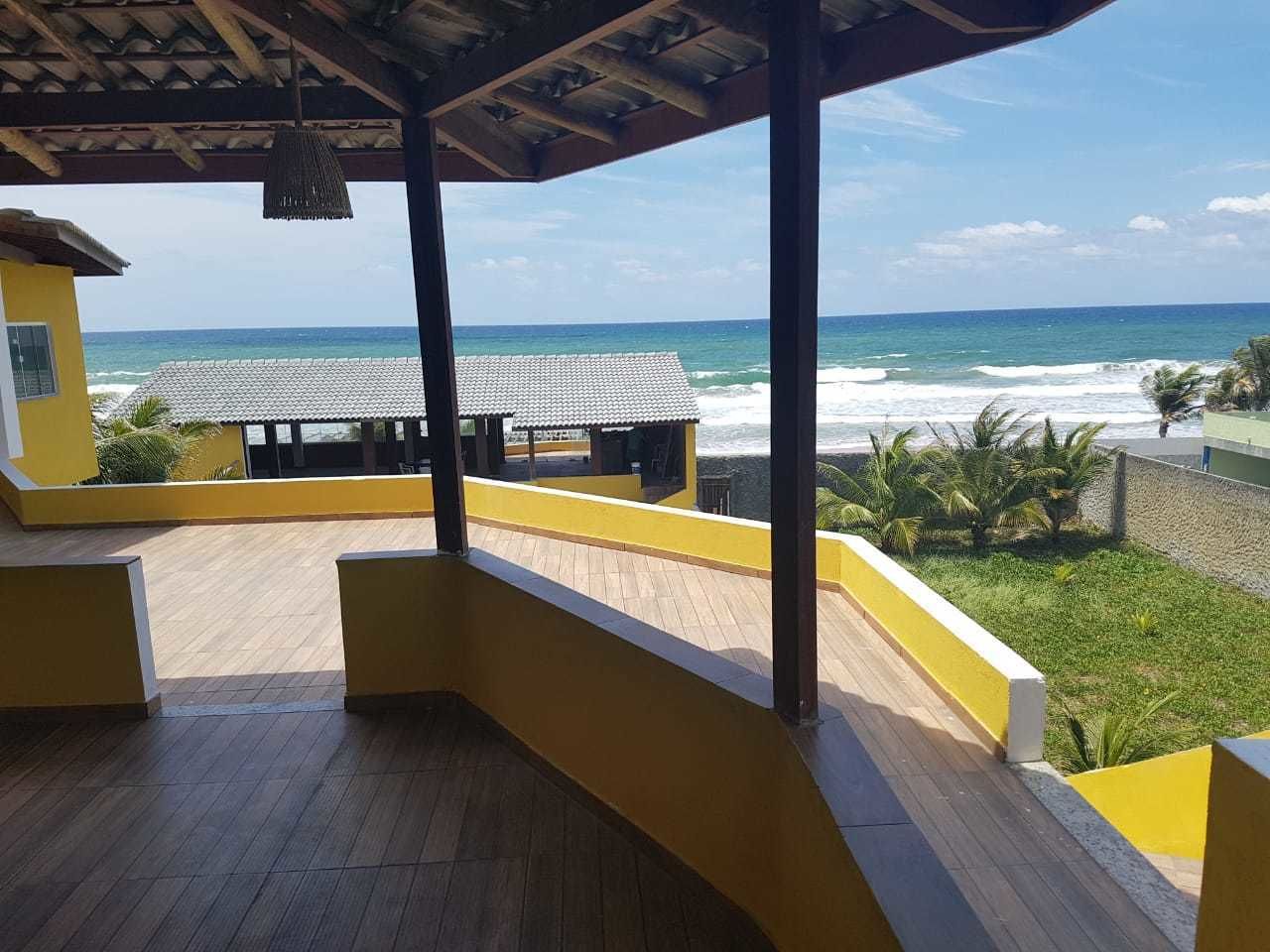 Un espace extérieur abrité, doté d’un plancher en bois et de murs jaunes, offrant une vue sur l’océan et la plage.