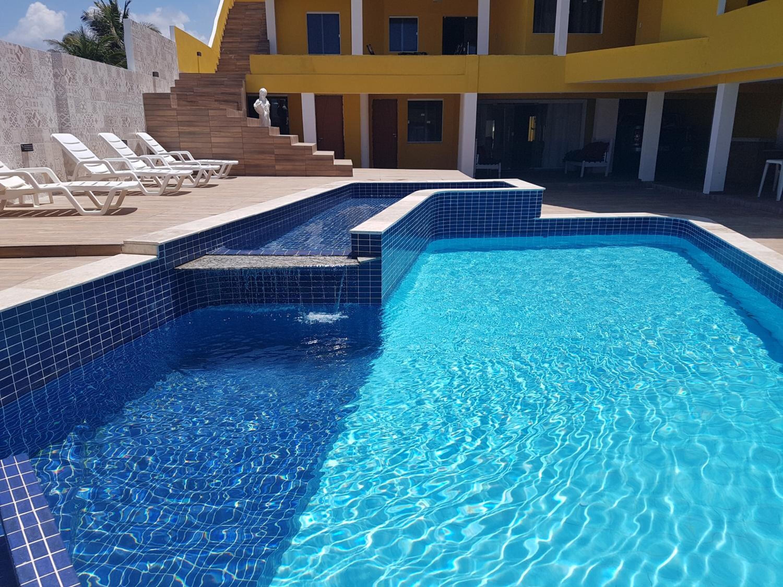 A swimming pool with blue tiles, a sun deck with lounge chairs, and a building with yellow walls and a staircase.