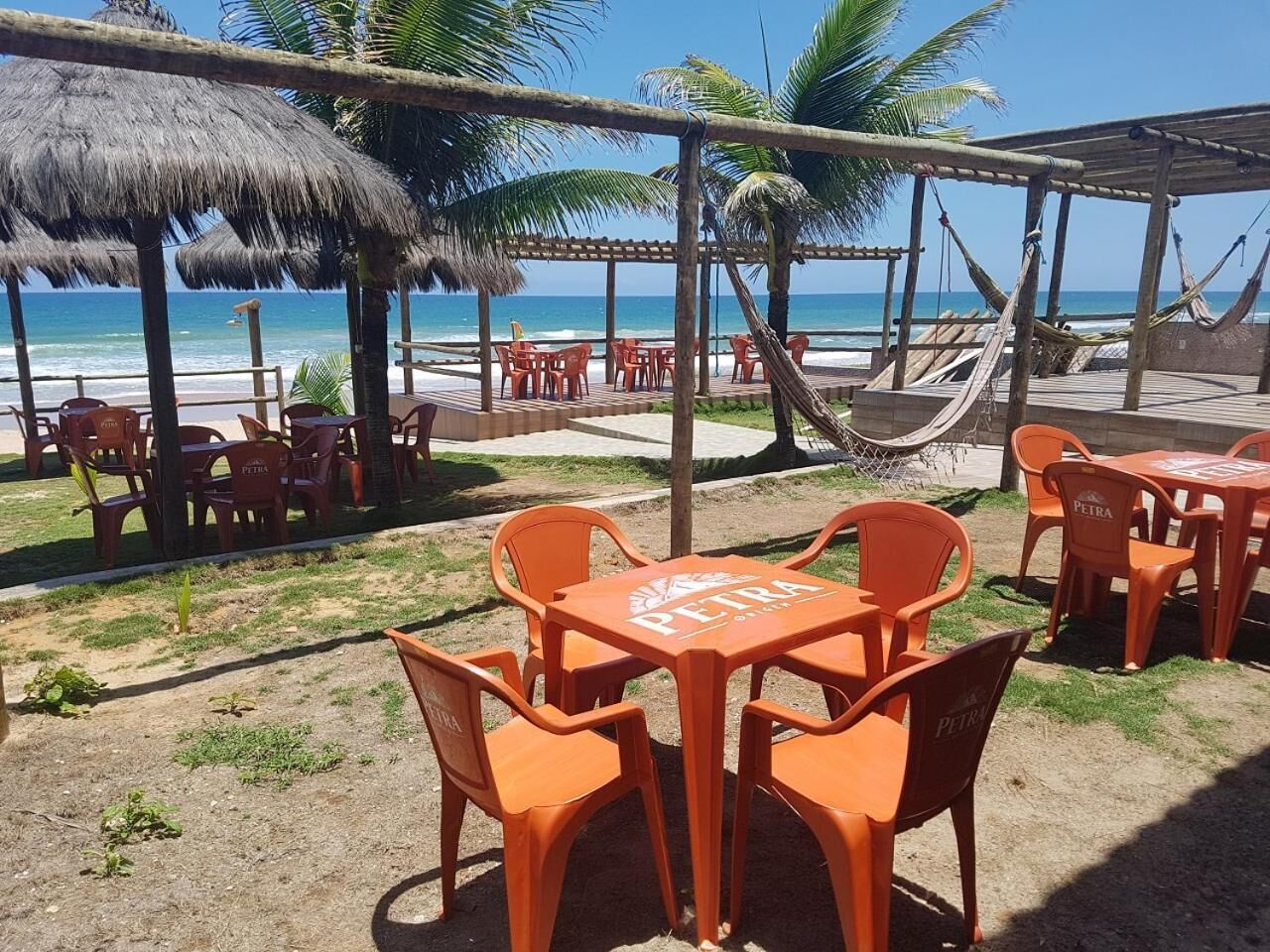 Un espace extérieur aménagé avec des chaises et des tables en plastique orange, une structure dotée d’un toit de chaume et offrant une vue sur l’océan.