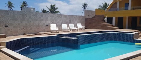A rectangular swimming pool with blue tiles, surrounded by a wooden deck and white lounge chairs.