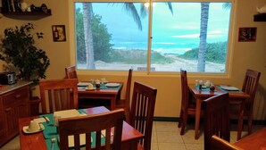 A dining area with wooden furniture, a large window with a view of the beach and ocean, and a shelf with books and kitchen items. - Drake Inn (Puerto Villamil)