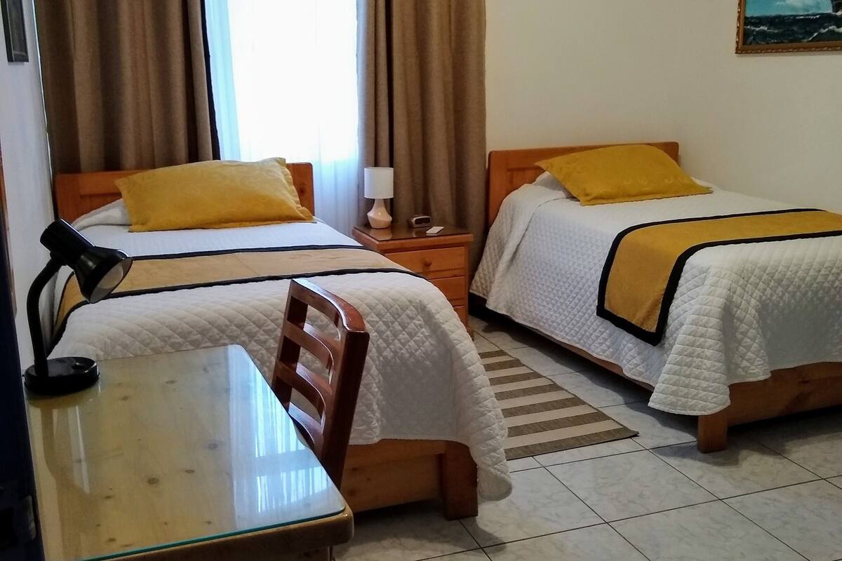 A hotel room with two beds, a desk, a chair, a nightstand, and a window with curtains.