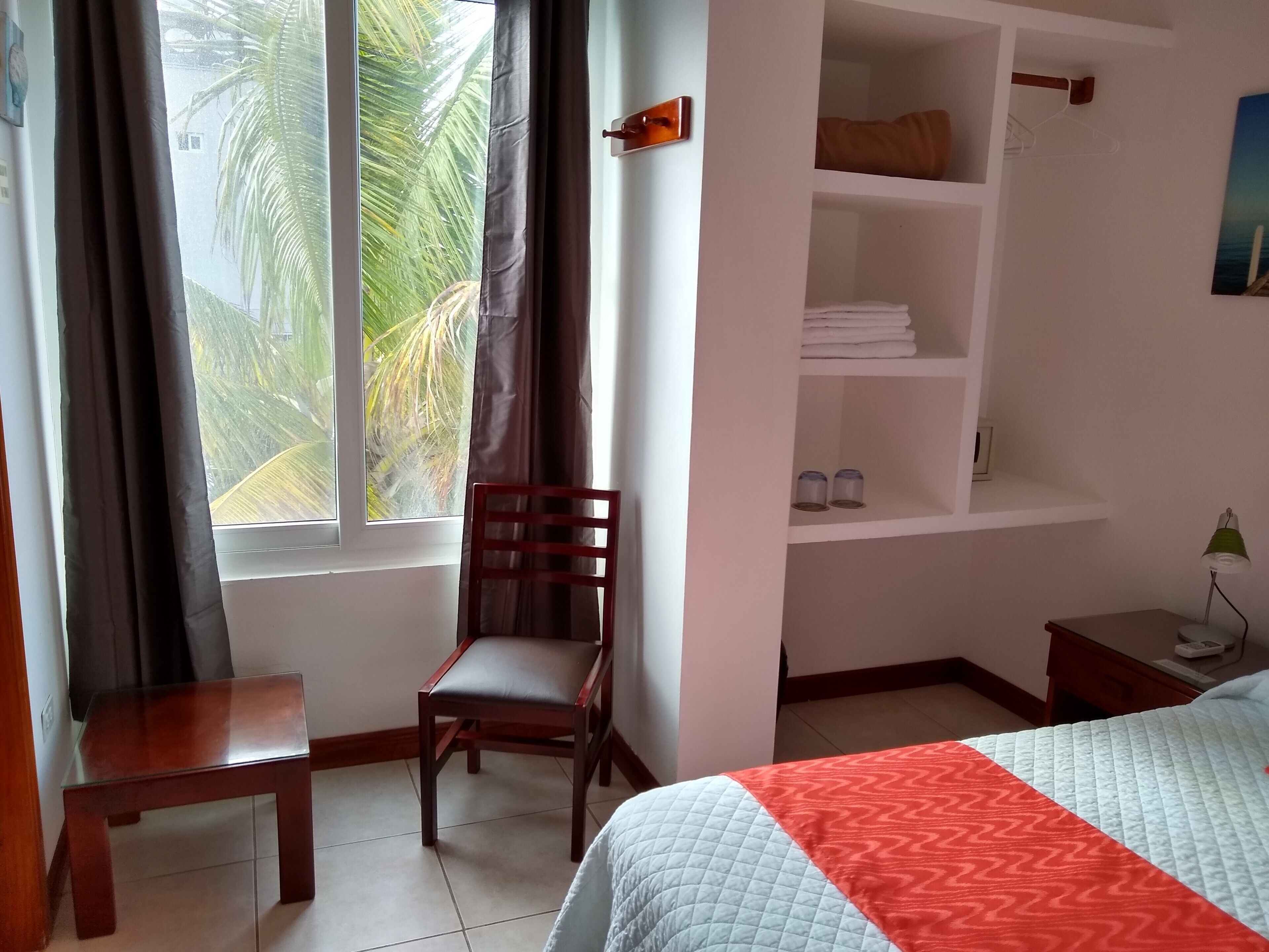 A hotel room with a bed, a chair, a small table, a shelf with folded towels, and a view of palm trees through the window.