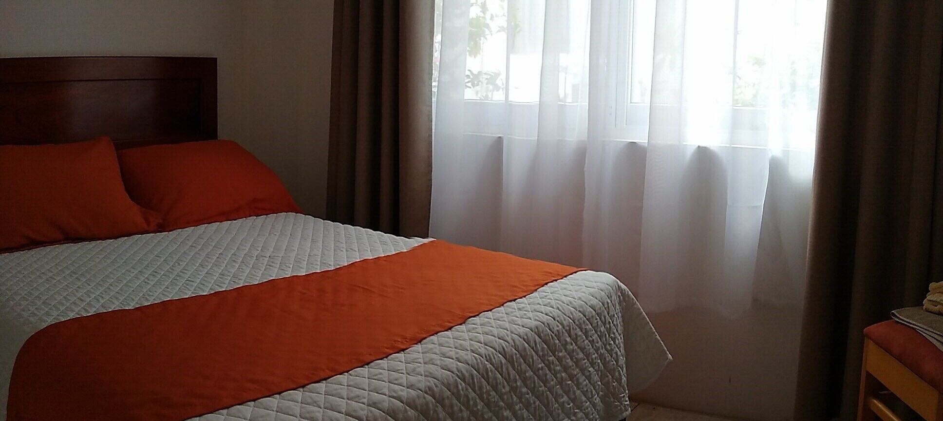 A bed with an orange and white quilt, a wooden headboard, and a window with sheer curtains.