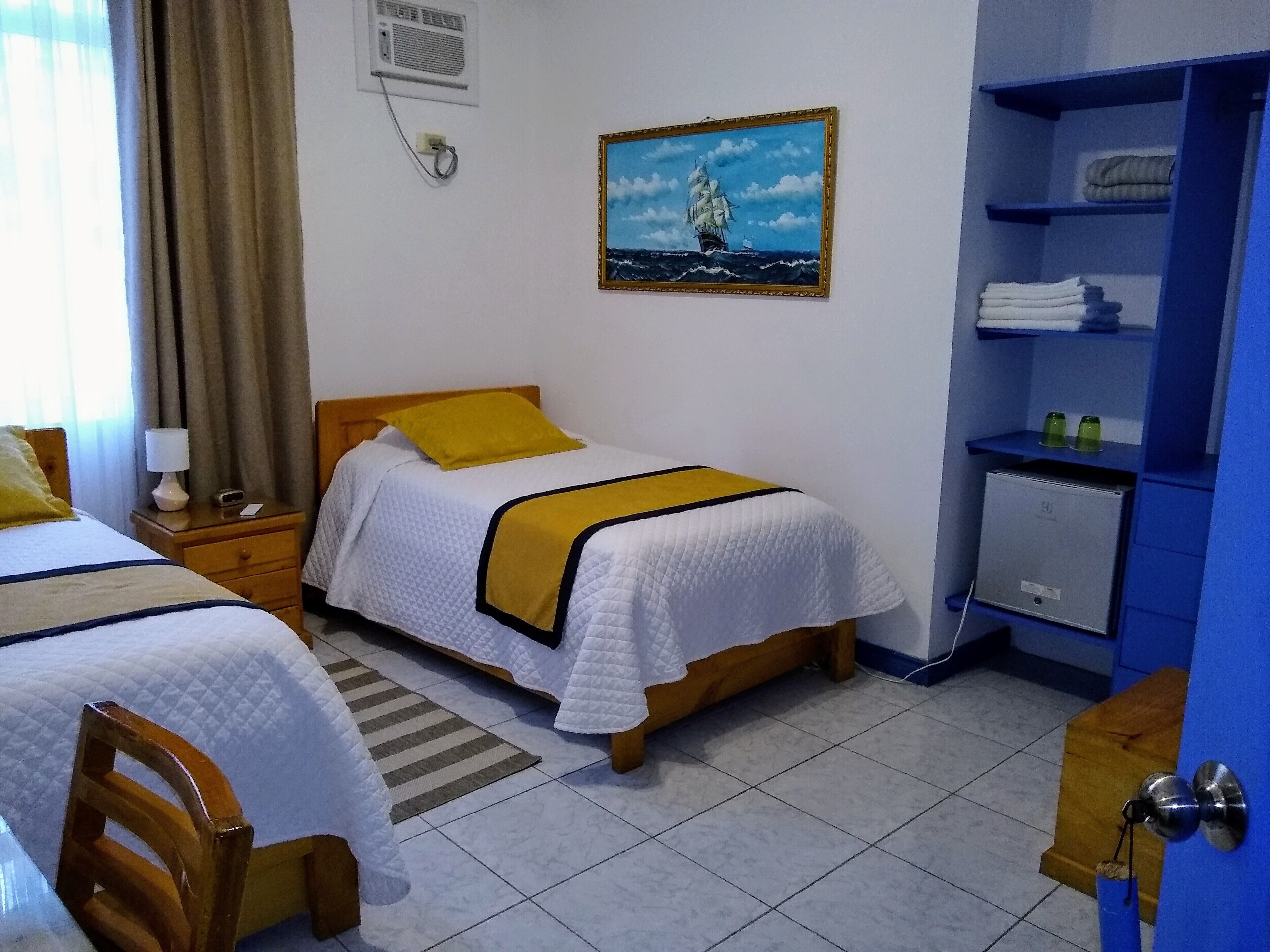 A hotel room with two beds, a painting of a sailboat, a desk with a lamp, and a shelf with towels.