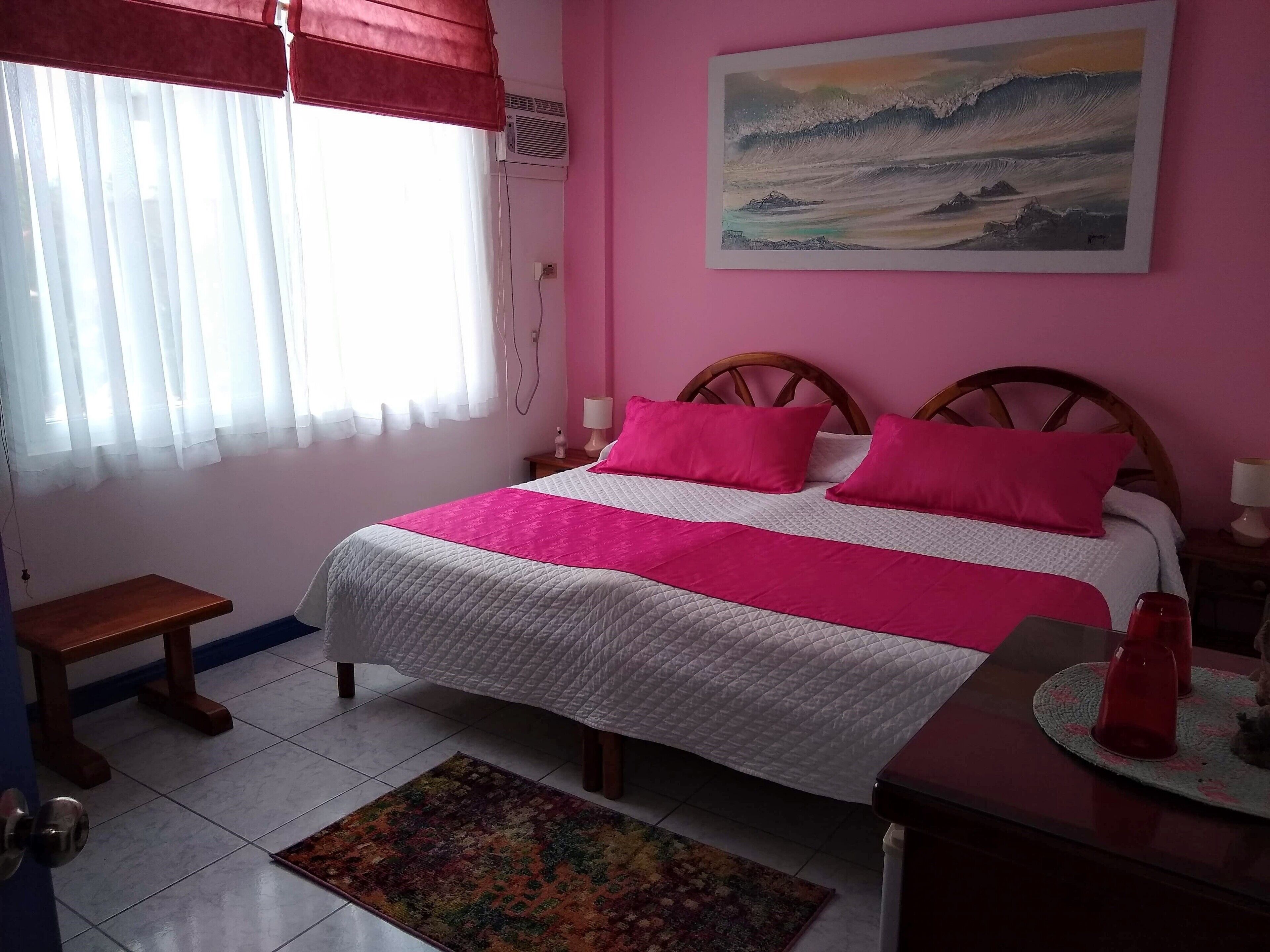 A bedroom with a bed, a bedside table, a nightstand, a window with curtains, a wall-mounted air conditioner, and a painting.
