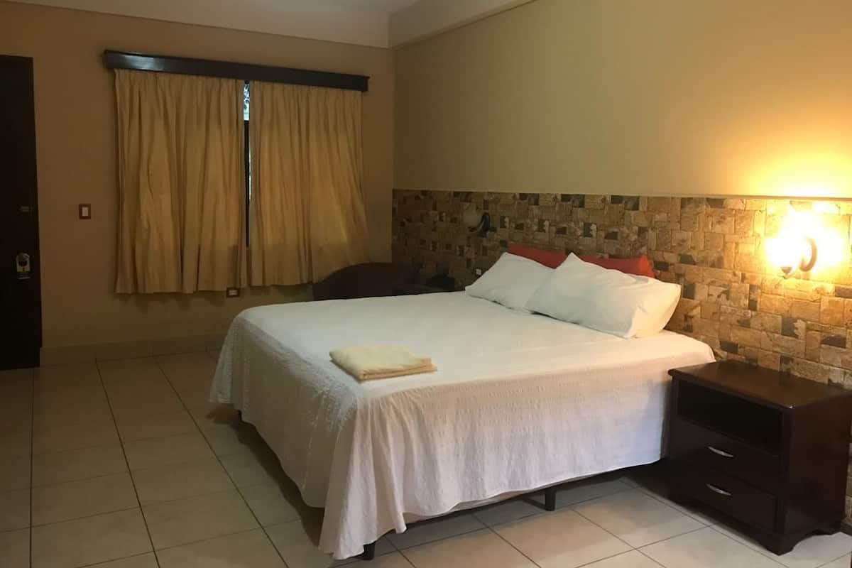 A hotel room with a bed, a nightstand, a wall-mounted light, and a window with curtains.