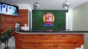 A hotel reception desk with a wooden finish, a green wall with a 'SJ Hotel San Jose' sign, and a television mounted above. - Hotel San José (San José)