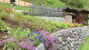 Een rustieke hut met een stenen muur en houten omheining, omringd door weelderige begroeiing en bloeiende planten.