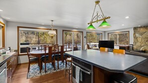 A modern kitchen with a large island, wooden chairs, and a dining table. There are large windows providing views of the outdoors. - Diamond on the Sky: Riverfront mountain lodge w/ views, AC & WiFi (Skykomish)