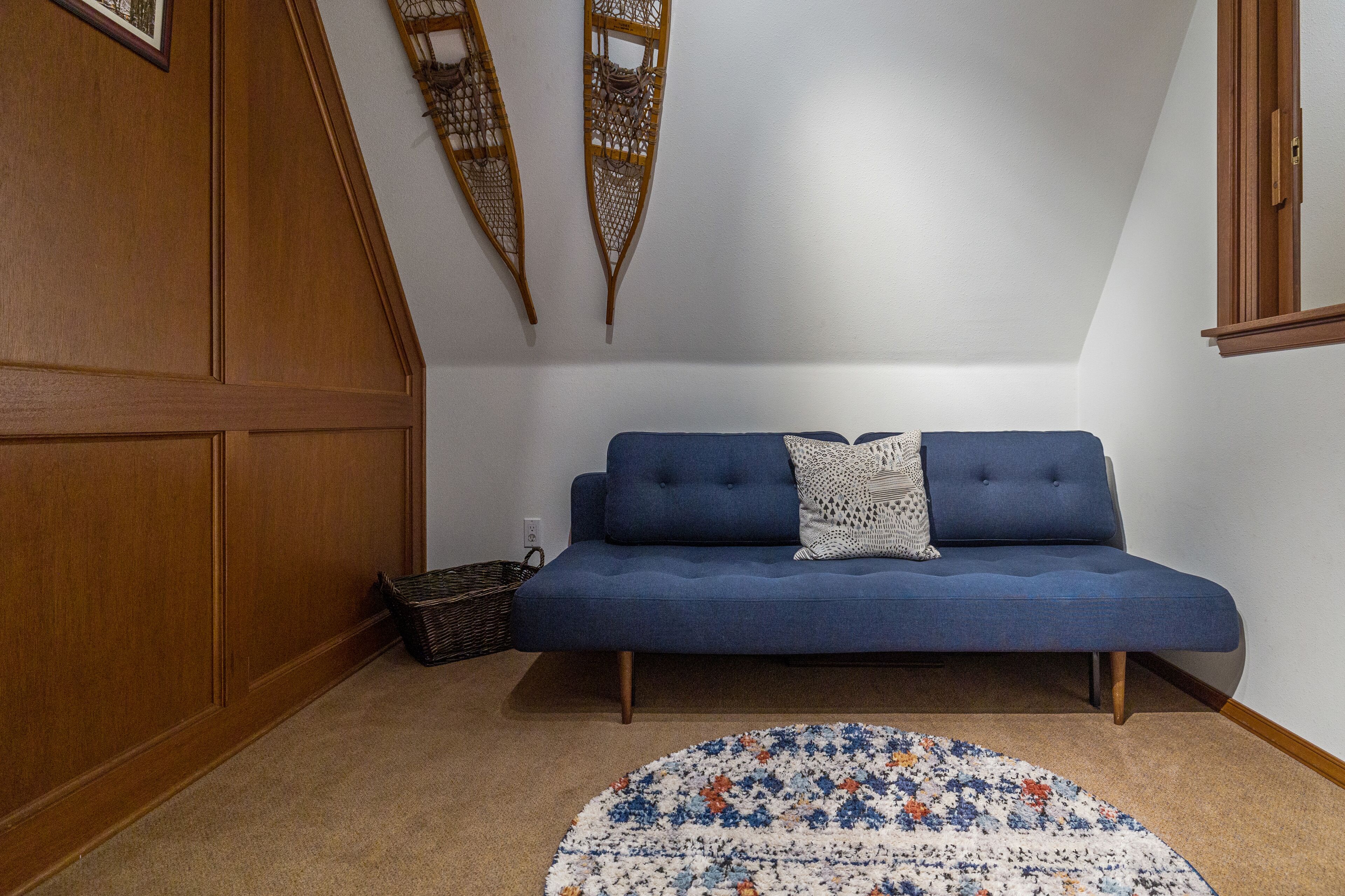 Un sofá azul con un cojín estampado, una cesta de mimbre y una alfombra floral en una habitación con paneles de madera y techo blanco.