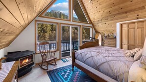 A cozy attic bedroom with a wooden ceiling, a large bed, a fireplace, and a view of a forested mountain through large windows. - Diamond on the Sky: Riverfront mountain lodge w/ views, AC & WiFi (Skykomish)