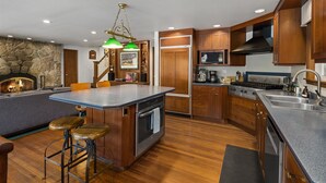 A kitchen with a stone wall, wooden cabinets, a central island with stools, a built-in oven, and a stove. - Diamond on the Sky: Riverfront mountain lodge w/ views, AC & WiFi (Skykomish)