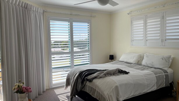 Ein Schlafzimmer mit Bett, Vorhängen, einem Fenster mit Jalousien, einem Deckenventilator und einem Nachttisch mit Lampe.