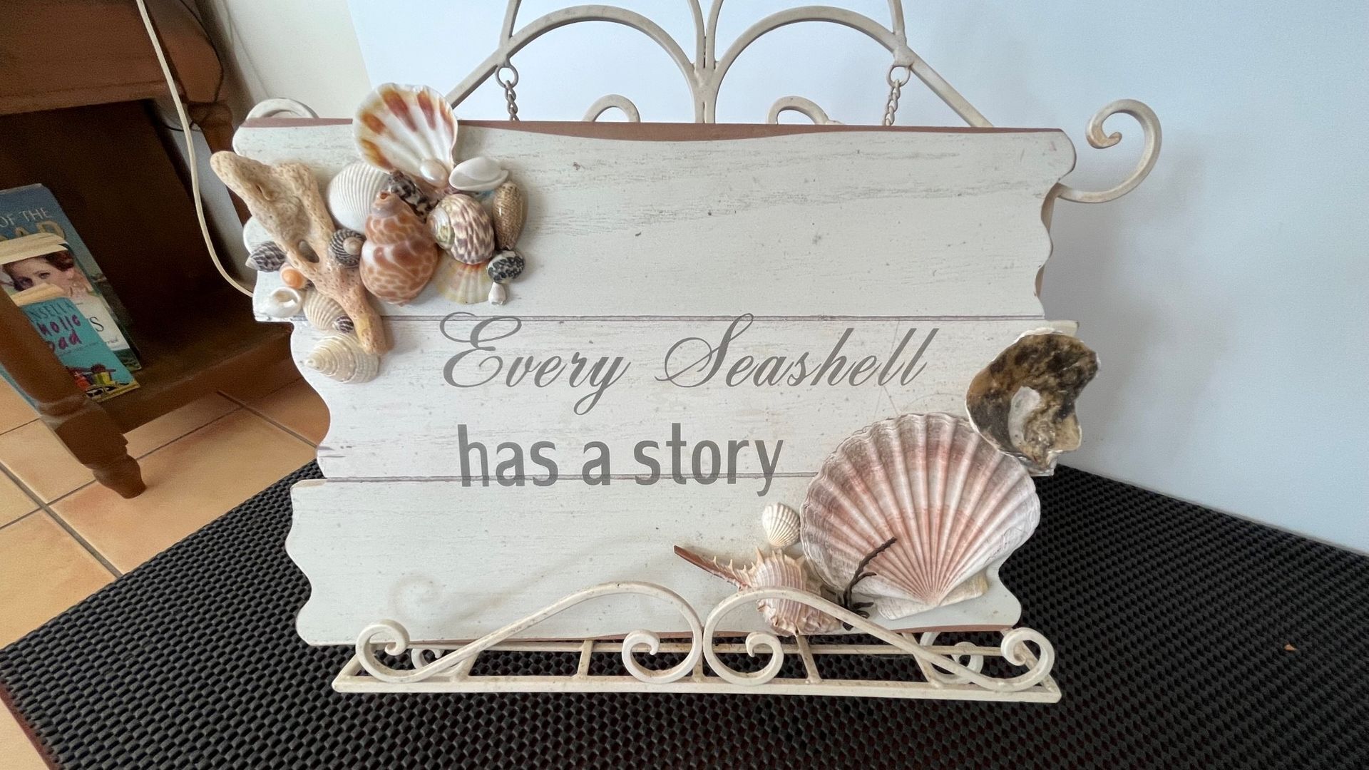 Ein dekoratives Schild mit Meeresschnecken und der Aufschrift "Every Seashell has a story" auf einer Holzfläche.