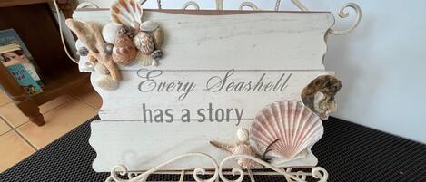 Ein dekoratives Schild mit Meeresschnecken und der Aufschrift "Every Seashell has a story" auf einer Holzfläche.
