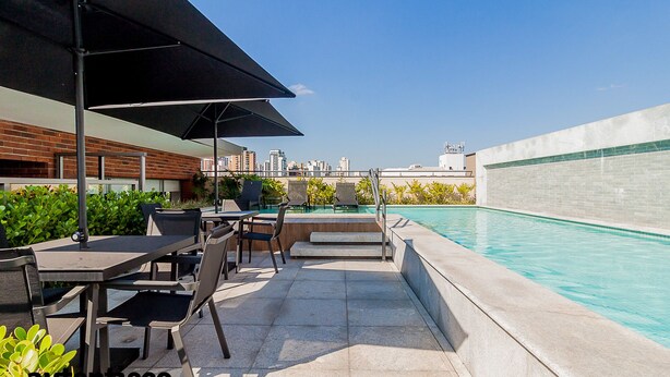 An outdoor pool area with a city view, including a poolside seating area with tables and chairs, and a covered section with umbrellas. - Autenticco - Tita Salzano (São Paulo)