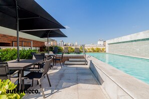 Un espace piscine extérieur offrant une vue sur la ville, comprenant un coin salon avec tables et chaises, ainsi qu’une zone abritée avec des parasols.
