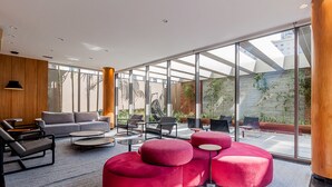 A modern interior with a grey sofa, red ottomans, and a glass wall offering a view of a brick wall and greenery. - Autenticco - Tita Salzano (São Paulo)