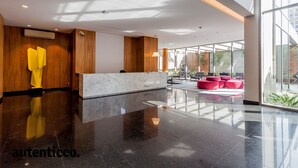 A modern lobby with a marble reception desk, wooden accents, and a glass wall offering a view of outdoor seating. - Autenticco - Tita Salzano (São Paulo)