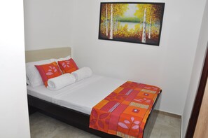 Ein ordentlich bezogenes Bett mit weißen Leinentüchern, orangen Blumenkissen und einer farbenfrohen Decke, sowie ein Baumgemälde an der Wand.