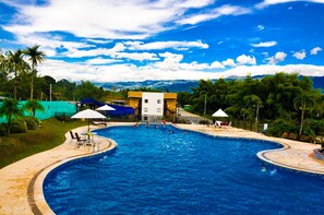 Ein großer Pool mit gewölbter Kante, umgeben von einer gepflasterten Fläche mit Sonnenschirmen und Liegestühlen.