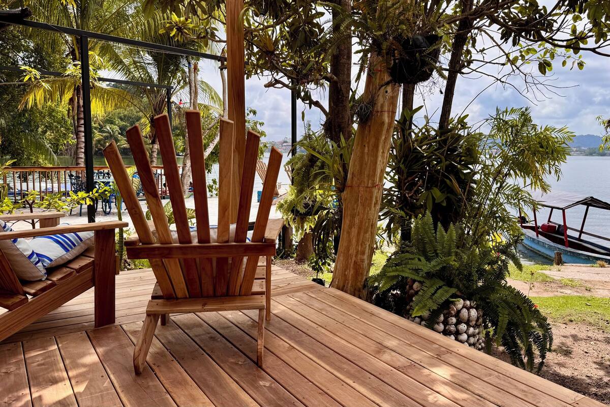 Um deck de madeira com uma cadeira e um sofá, com vista para um lago onde um barco está atracado.