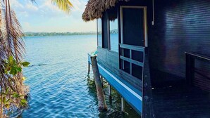 Un bungalow au toit de chaume doté d’une terrasse en bois qui s’étend au-dessus de l’eau, entouré de palmiers.