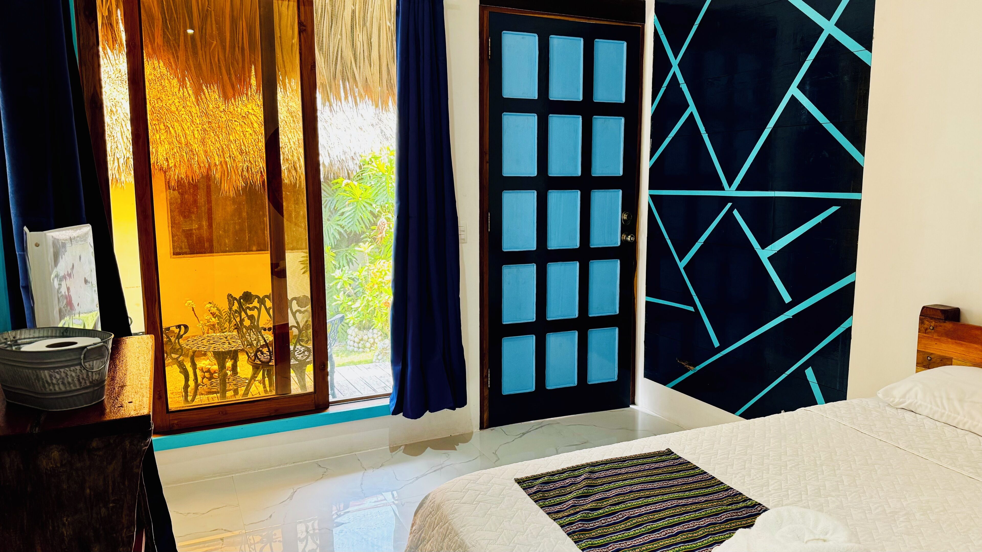 Una habitación con cama, un escritorio con lámpara, una ventana con cortinas y una puerta con diseños geométricos.