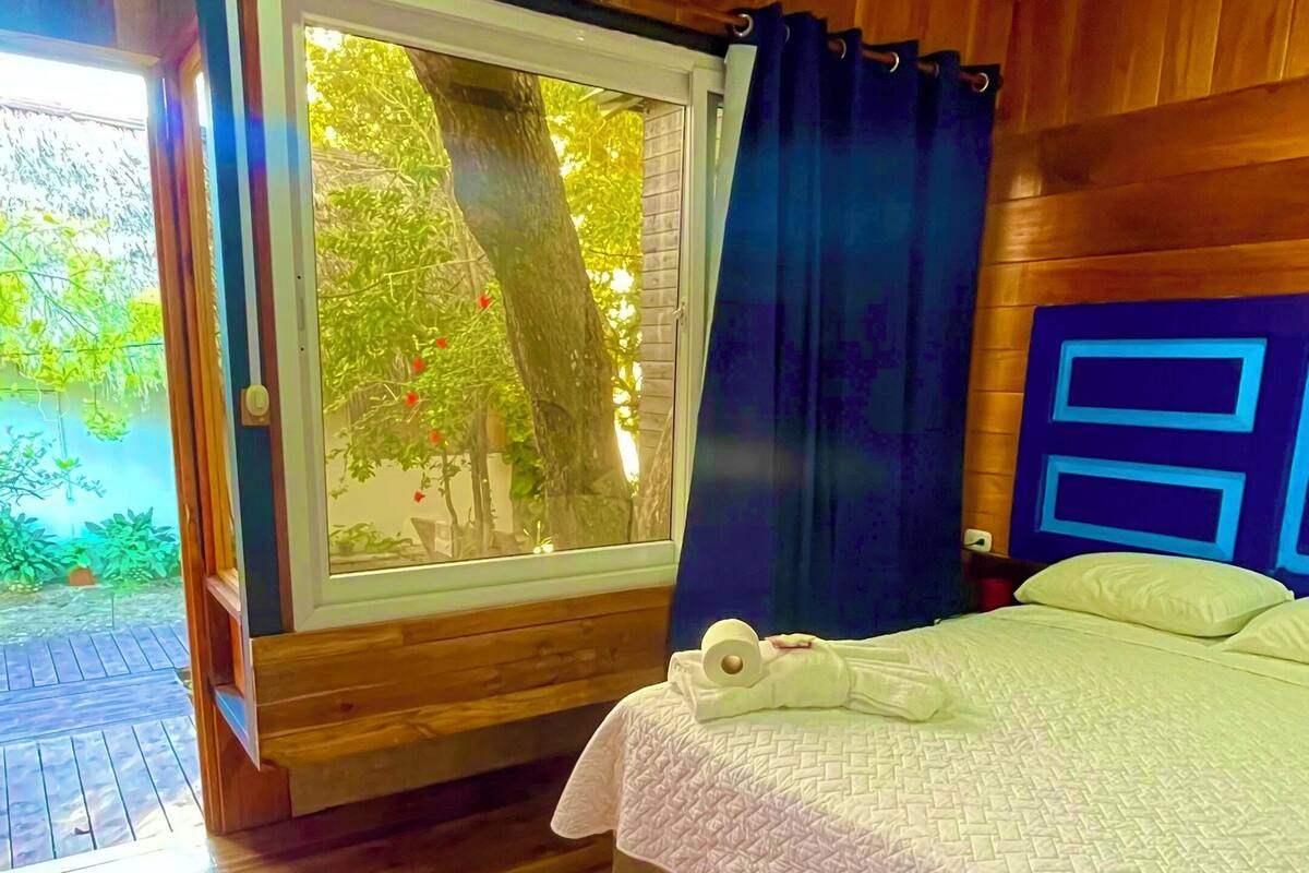 Quarto com piso de madeira, cama com colcha branca, janela com vista para vegetação e cabeceira azul.