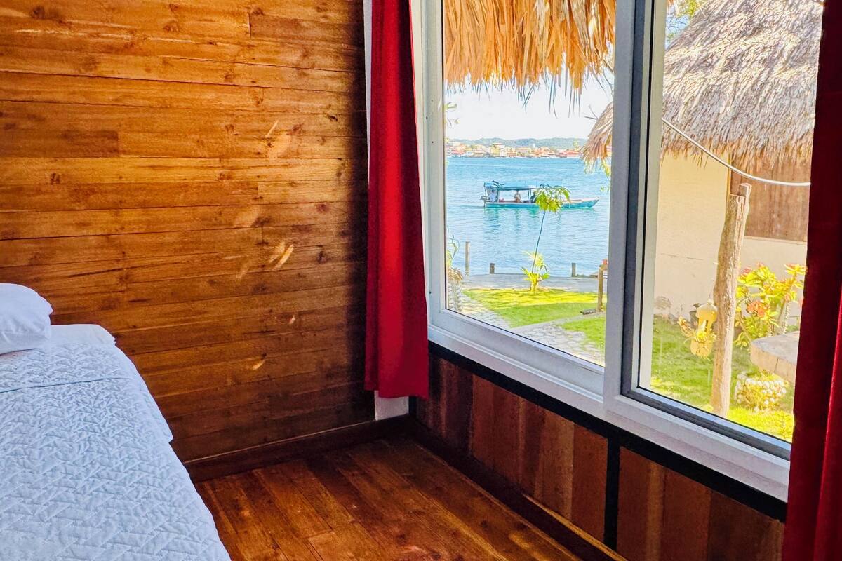 Quarto de madeira com cama, janela com cortinas vermelhas e vista para um corpo d'água e edifícios.