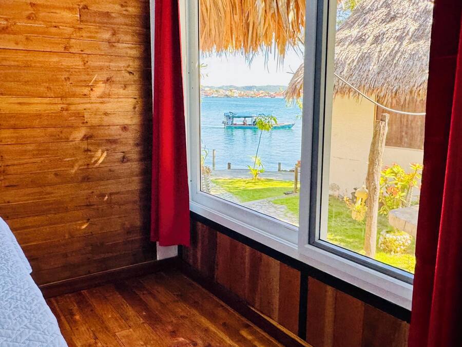 A wooden room with a bed, a window with red curtains, and a view of a water body and buildings.