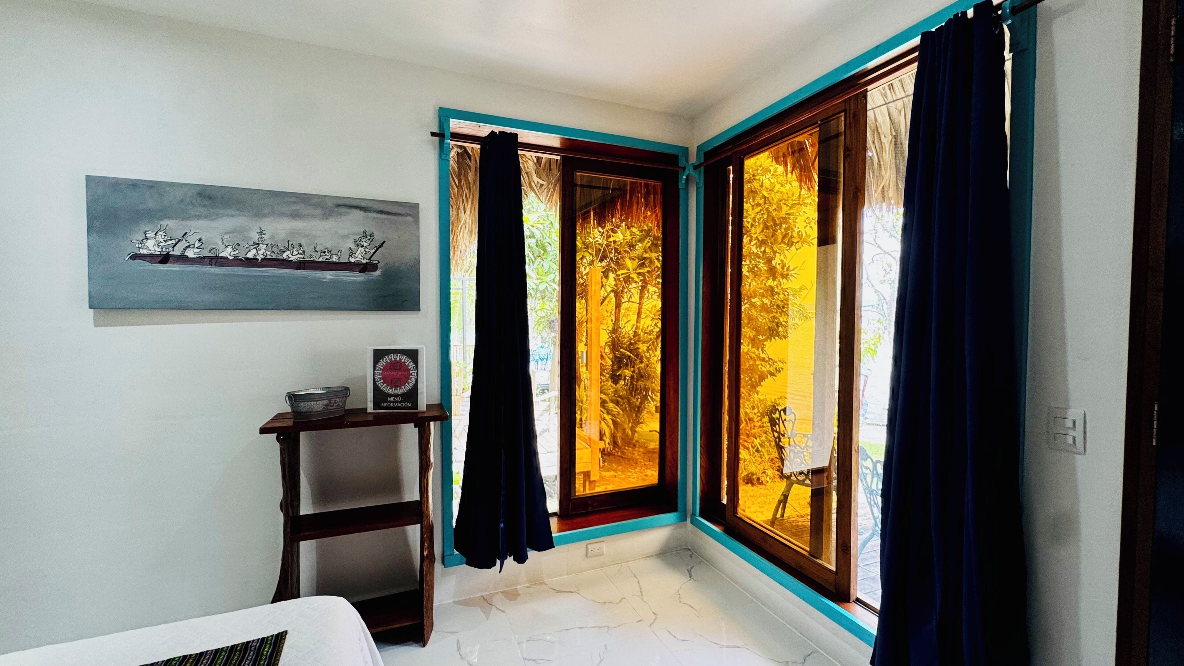 A corner room with a bed, a small table with a bowl, a framed picture, and a window with curtains.