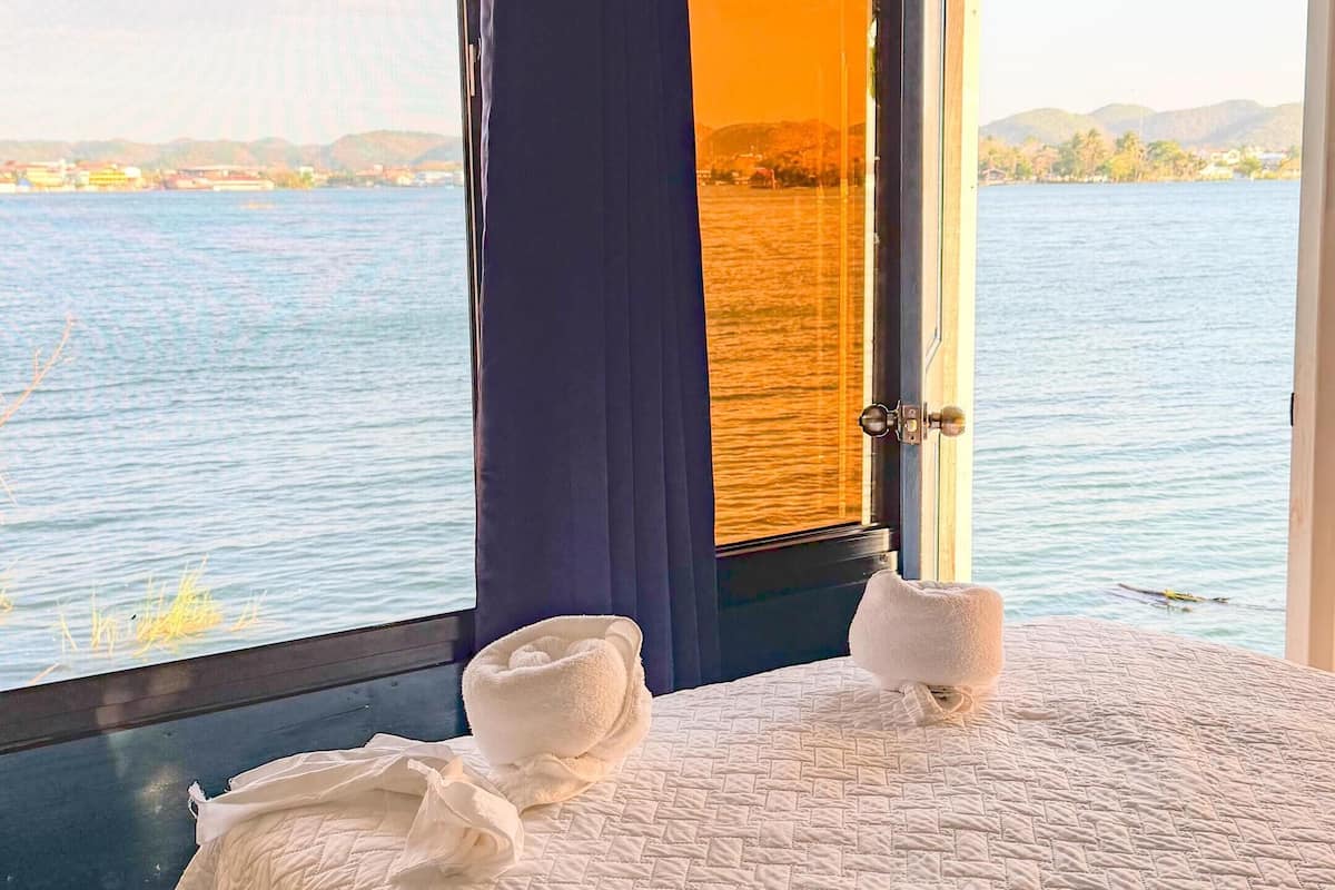 Cama com colcha branca, duas toalhas brancas e vista para um corpo d'água através de janela com moldura de madeira e telhado de palha.