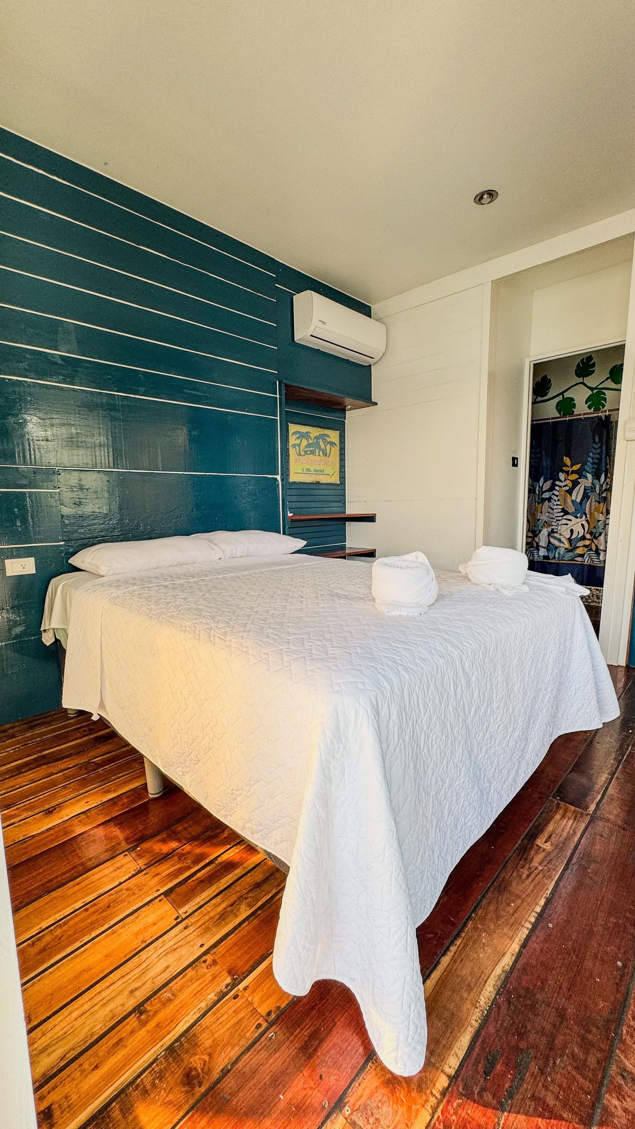 A single bed with a white bedspread in a room with wooden flooring and a teal-colored wall.