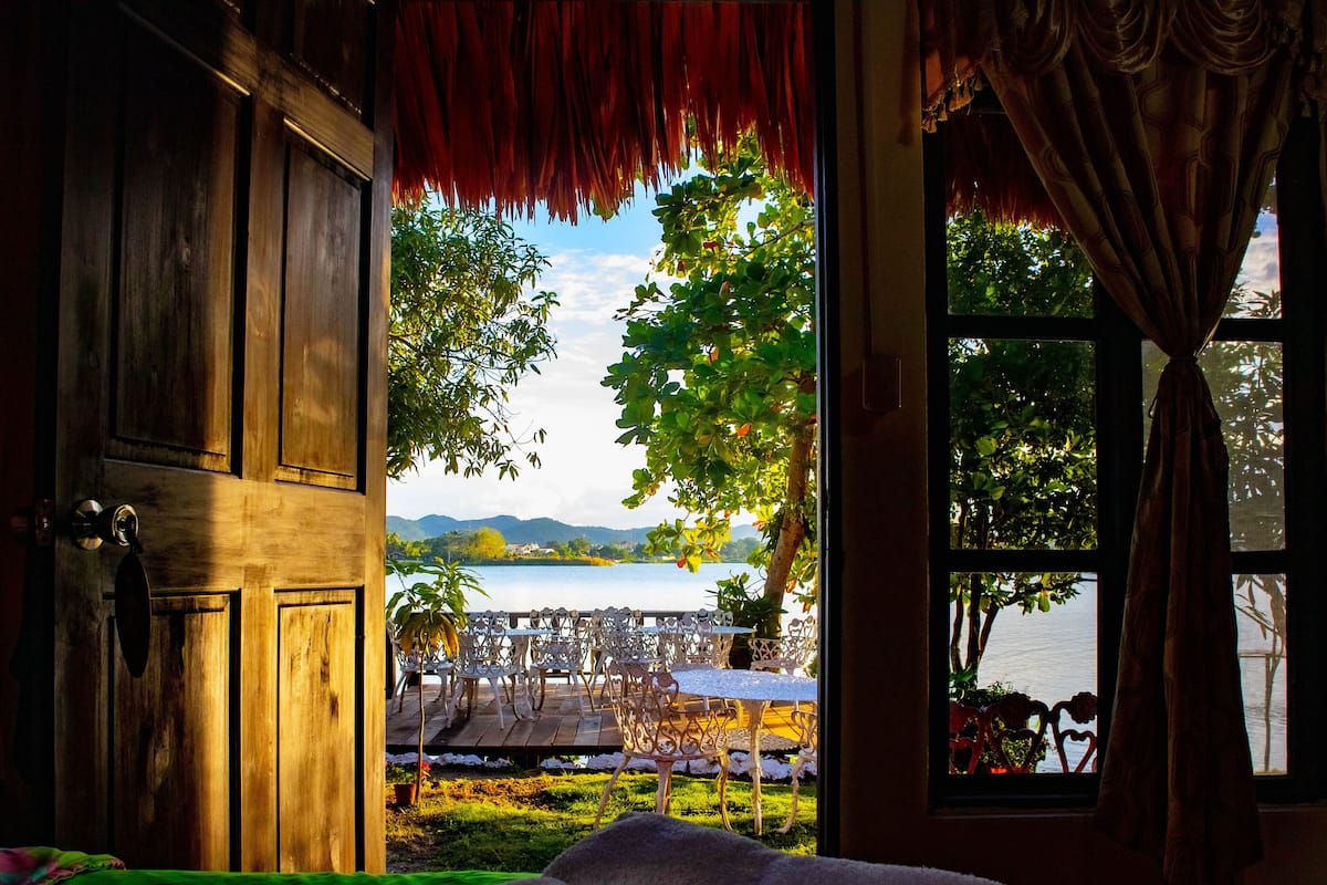 Quarto com cama, janela com vista para um lago e montanhas, e porta de madeira.