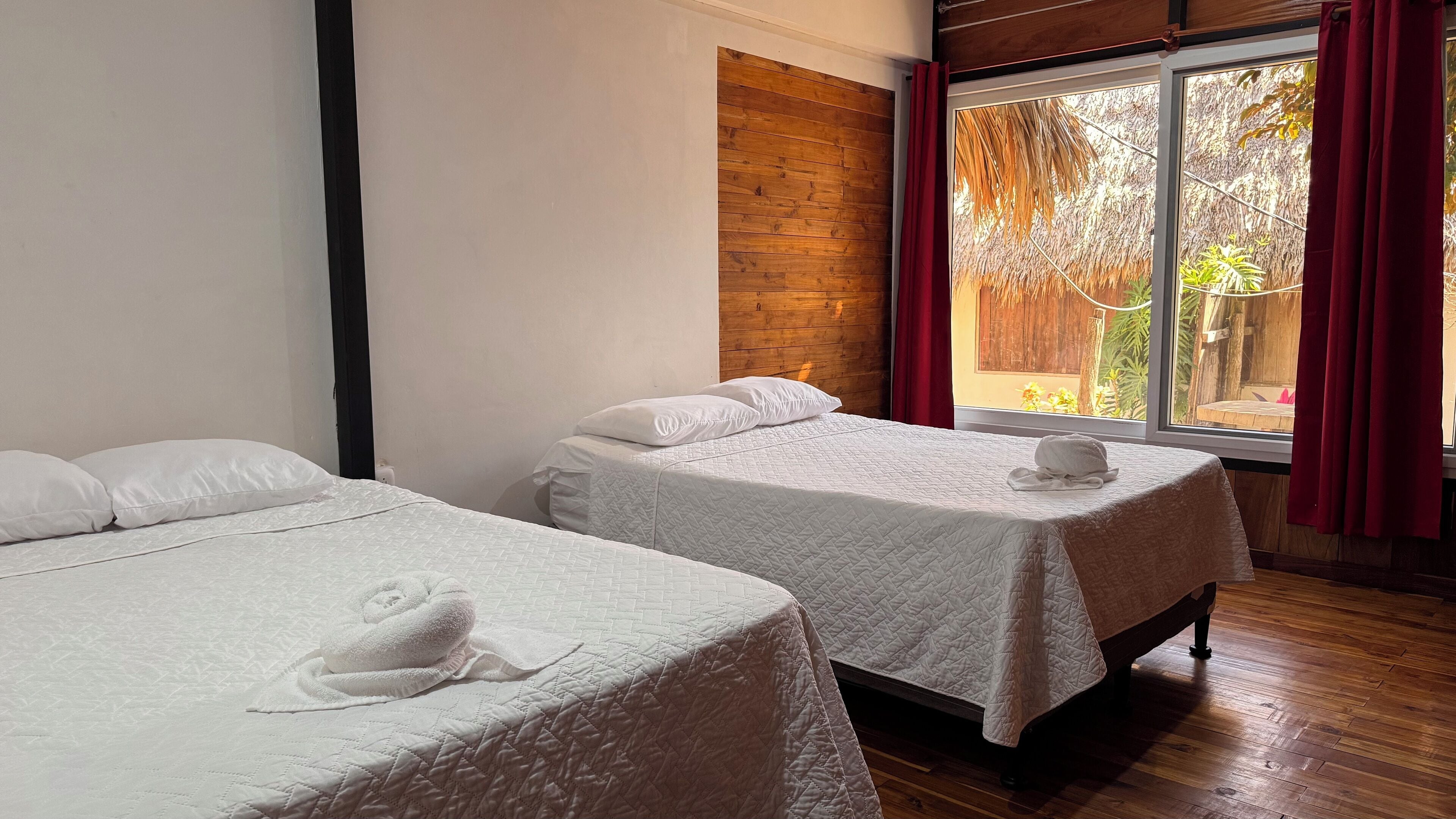 A hotel room with two beds, a wooden wall, and a window with a view of a garden.