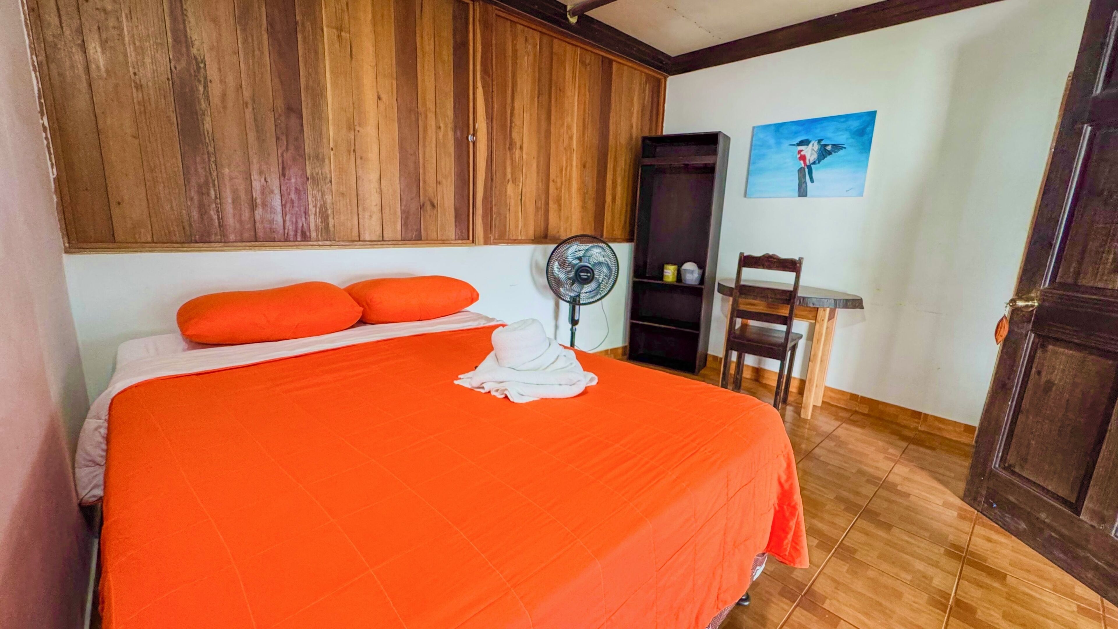 Un dormitorio con una cama grande, una colcha naranja, una toalla blanca, un armario de madera, un ventilador, una silla y un cuadro en la pared.