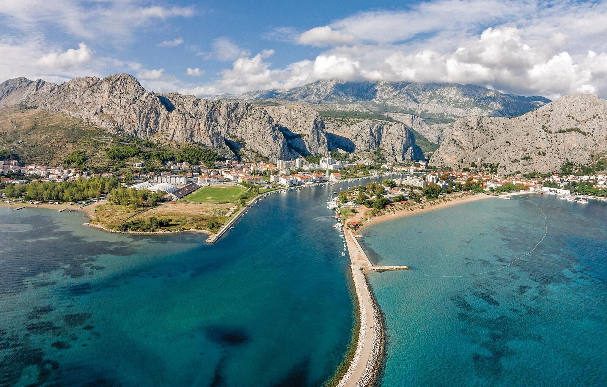 Vista aerea di una cittadina costiera con una marcata catena montuosa sullo sfondo, un porto turistico con barche e un mare blu cristallino.