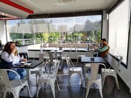 Un café al aire libre con mesas y sillas, amplias ventanas con vistas a zonas verdes y una decoración interior moderna.