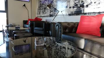 A modern living room with a glass coffee table, black leather sofa with red cushions, a mounted TV, and a cityscape wall mural.