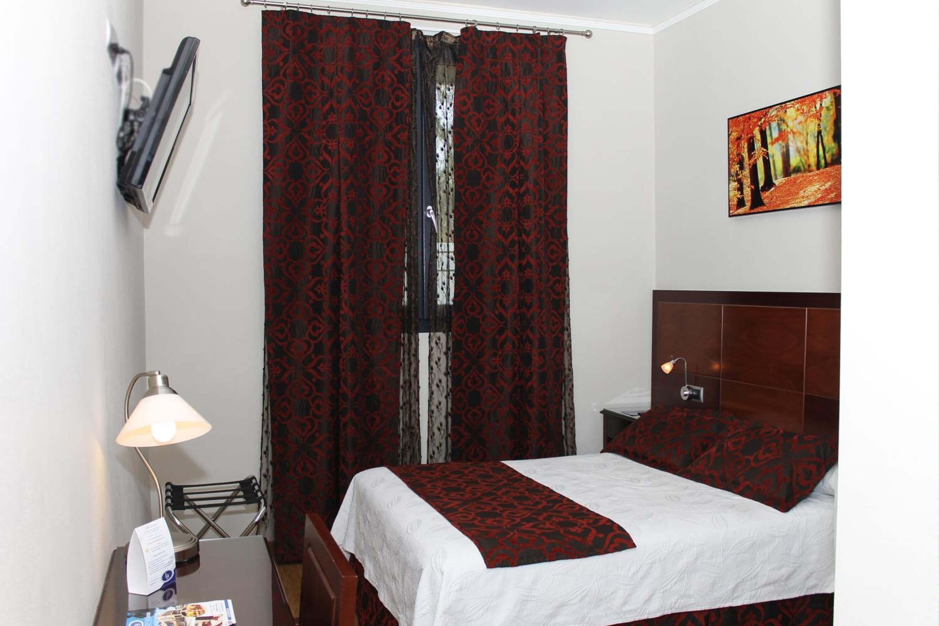 A hotel room with a bed, a nightstand, a lamp, a window with red patterned curtains, and a painting on the wall.