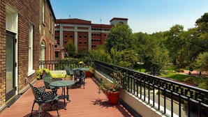 A balcony with outdoor seating, a brick building, and a view of a campus with trees and a building in the background.