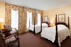 A hotel room with two beds, floral curtains, a wooden chair, a bedside table with a lamp, and framed artwork on the wall. - Planters Inn (Savannah)