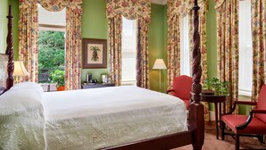 A bedroom with a large bed, floral curtains, a wooden chair, a table with a lamp, and a framed picture on the wall.