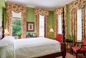 Een slaapkamer met een groot bed, bloemen gordijnen, een houten stoel, een tafel met een lamp en een lijst met een foto aan de muur.