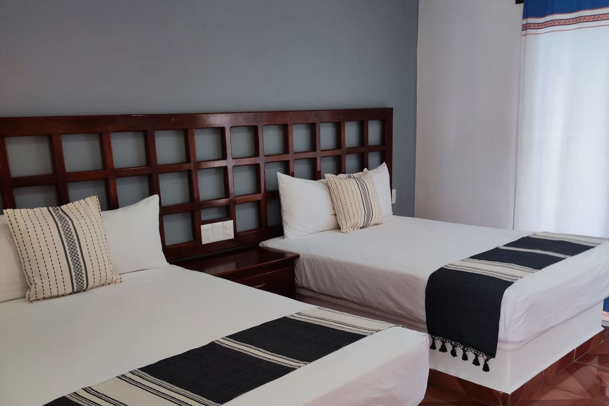 Habitación de hotel con dos camas, cabecero de madera, ropa de cama blanca, manta oscura y ventana con cortina azul.