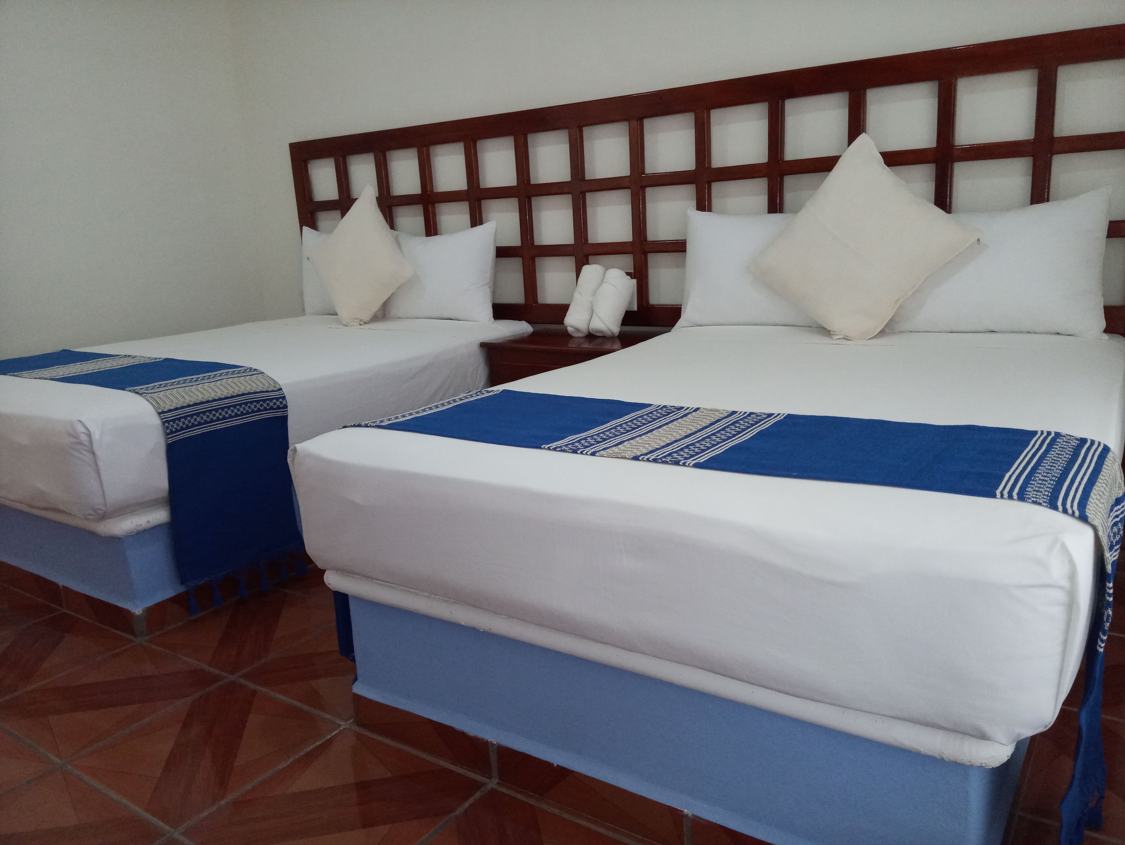 A hotel room with two beds, each with white bedding and blue accents, a wooden headboard, and a tiled floor.