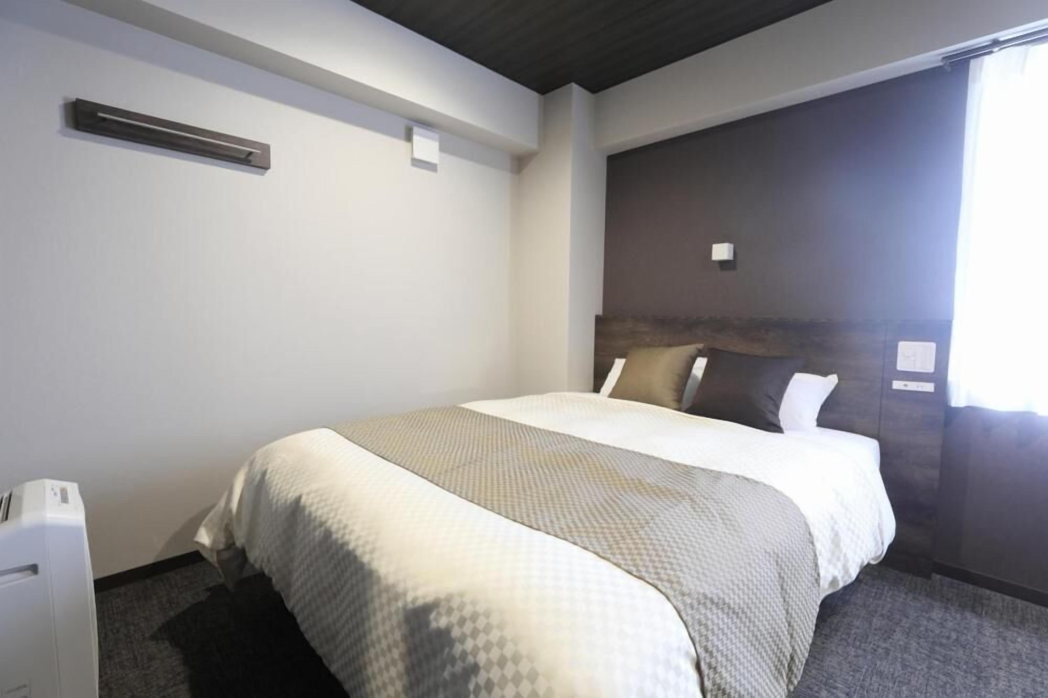 A hotel room with a bed, a bedside table, a headboard, and a window with curtains.